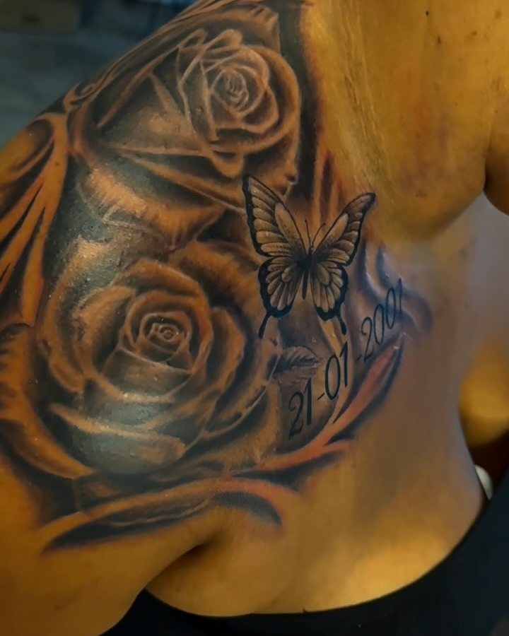 Black and grey roses with a butterfly and a date tattooed on a shoulder